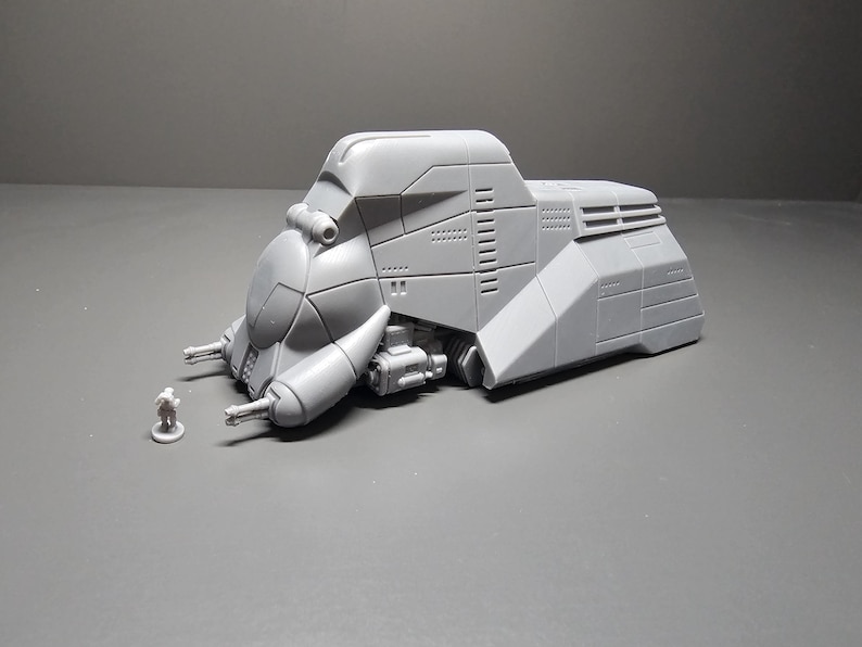 May include: A gray, futuristic vehicle model with a detailed design, featuring a rounded front, side-mounted protrusions, and a small figure for scale. The model has a textured surface and is set against a gray backdrop.