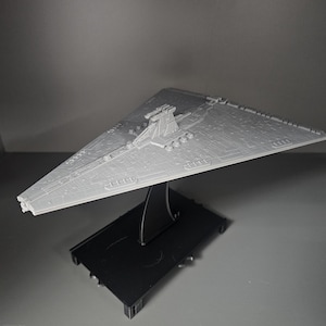 May include: A grey model of a Star Destroyer spaceship, displayed on a black stand. The spaceship has a triangular shape with detailed surface features. The stand is rectangular with a curved support.