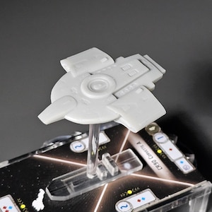 May include: A white plastic spaceship model with a circular centre and a raised section on the top. The model is mounted on a clear plastic stand and is displayed on a black and white game board with the text "CR90 Corvette 4".