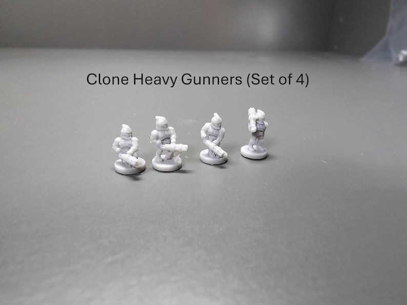 May include: Four small, grey Clone Heavy Gunners figurines. Each figure is posed with a weapon, standing on a small, round base. The text "Clone Heavy Gunners (Set of 4)" is above the figurines.