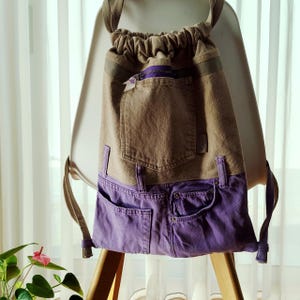 Op de afbeelding: Een rugzak met trekkoord gemaakt van gerecycled denim. Het bovenste gedeelte is een bruinachtige stof en de onderkant is paarse denim met zakken. De rugzak heeft een paarse rits en verstelbare bandjes.