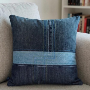 Puede incluir: Un cojín cuadrado hecho de tela de mezclilla azul índigo oscuro con una banda horizontal de mezclilla azul claro en el centro. El cojín está decorado con costuras verticales y horizontales en tonos de hilo azul, blanco y verde lima.