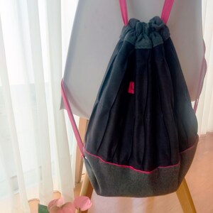 Könnte beinhalten: Ein dunkelgrauer Rucksack mit Kordelzug und rosa Akzenten. Die Tasche hat einen grauen Boden und eine graue Oberseite mit rosa Besatz und Trägern. Ein kleines rosa Quadrat ist auf der Vorderseite der Tasche sichtbar. Die Tasche hängt an einer weißen Oberfläche.