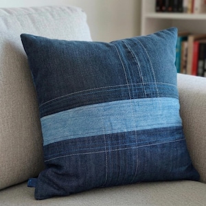Könnte beinhalten: Ein quadratisches Dekokissen aus Denim-Stoffbahnen in verschiedenen Blautönen. Das Design zeigt ein horizontales hellblaues Band in der Mitte, das durch dunklere indigoblaue Denim-Abschnitte oben und unten kontrastiert wird und mit sichtbaren Nähten versehen ist.