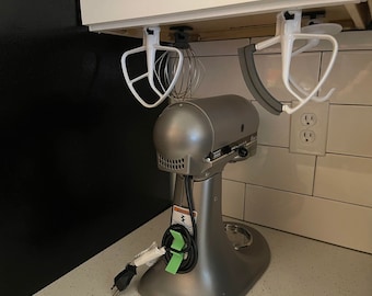 Op de afbeelding: Een zilveren keukenmixer met een wit snoer en groene elektrische tape op het snoer. De mixer staat op een wit aanrecht. Er hangen twee witte keukenutensilienhouders aan het kastje boven de mixer.