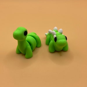 K&ouml;nnte beinhalten: Zwei gr&uuml;ne Plastik-Dinosaurier. Einer ist ein langer, segmentierter Dinosaurier mit schwarzen Augen. Der andere ist ein k&uuml;rzerer, runderer Dinosaurier mit wei&szlig;en Stacheln und schwarzen Augen.