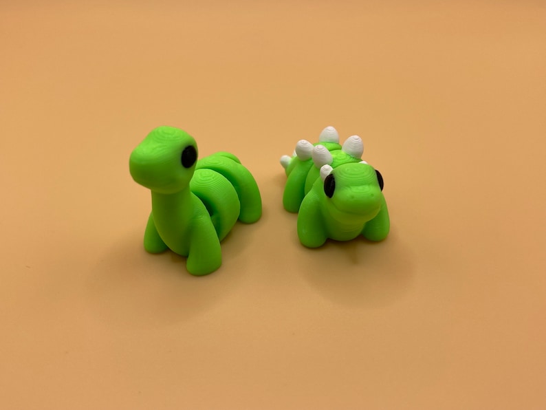 K&ouml;nnte beinhalten: Zwei gr&uuml;ne Plastik-Dinosaurier. Einer ist ein langer, segmentierter Dinosaurier mit schwarzen Augen. Der andere ist ein k&uuml;rzerer, runderer Dinosaurier mit wei&szlig;en Stacheln und schwarzen Augen.