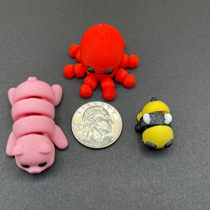 K&ouml;nnte beinhalten: Drei kleine 3D-gedruckte Figuren: ein roter Oktopus, ein rosa Schwein und eine gelbe und schwarze Biene. Die Figuren sind alle etwa so gro&szlig; wie eine 20-Cent-M&uuml;nze.