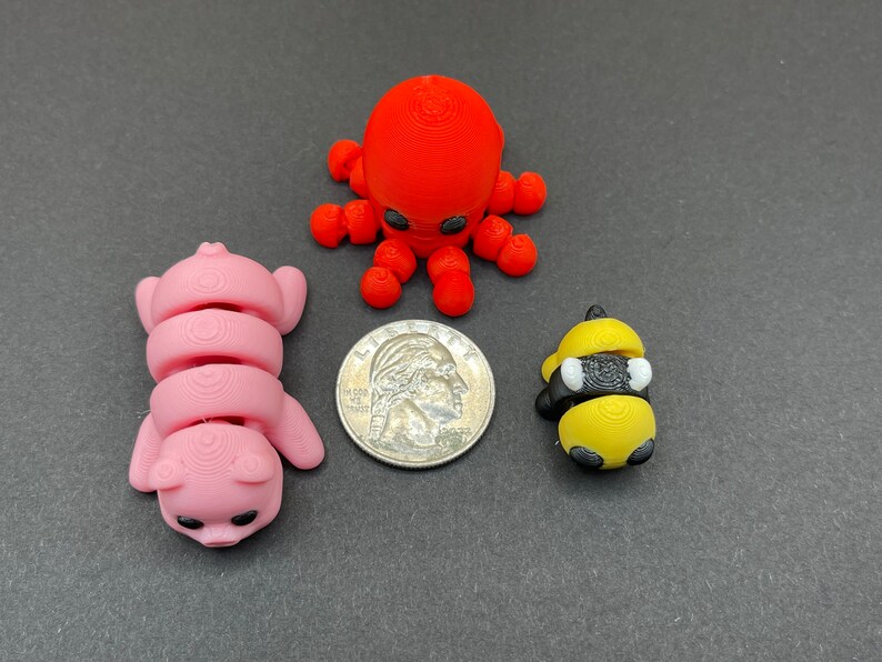 K&ouml;nnte beinhalten: Drei kleine 3D-gedruckte Figuren: ein roter Oktopus, ein rosa Schwein und eine gelbe und schwarze Biene. Die Figuren sind alle etwa so gro&szlig; wie eine 20-Cent-M&uuml;nze.