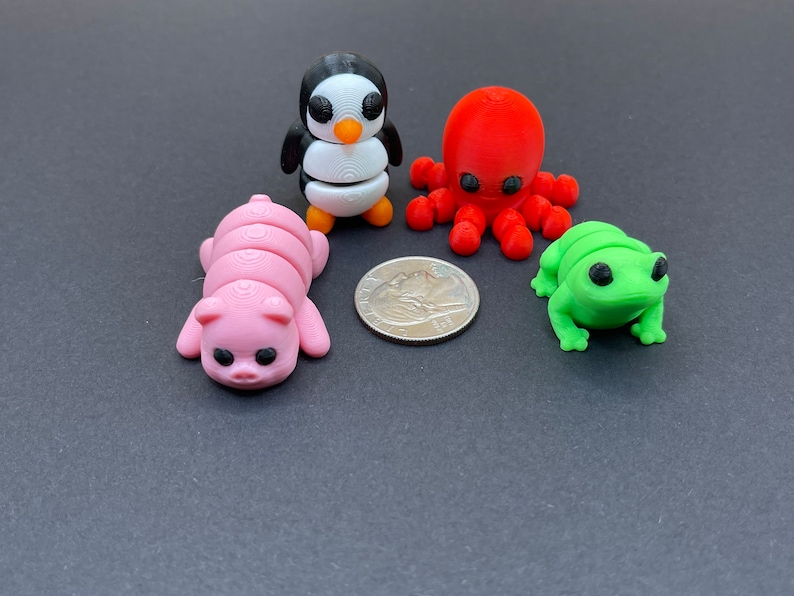 K&ouml;nnte beinhalten: Ein Set aus vier kleinen, bunten, 3D-gedruckten Tierfiguren: ein rosa Schwein, ein schwarz-wei&szlig;er Pinguin, ein roter Tintenfisch und ein gr&uuml;ner Frosch. Die Figuren sind etwa so gro&szlig; wie eine US-amerikanische 25-Cent-M&uuml;nze.