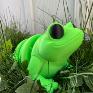 May include: A bright green frog figurine with large black eyes, sitting in a bed of green grass.