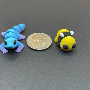 K&ouml;nnte beinhalten: Eine blaue und lila Eidechsenfigur und eine gelbe und schwarze Bienenfigur, beide etwa so gro&szlig; wie eine US-amerikanische 25-Cent-M&uuml;nze.