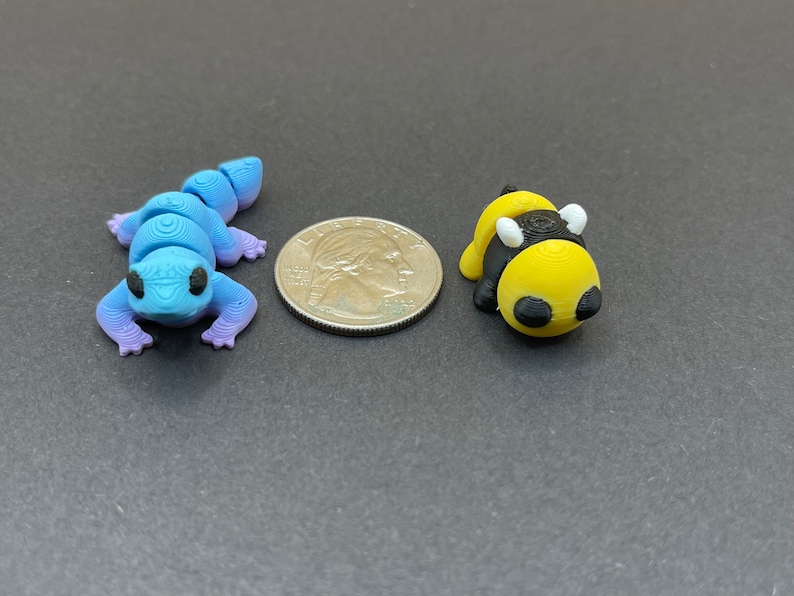 K&ouml;nnte beinhalten: Eine blaue und lila Eidechsenfigur und eine gelbe und schwarze Bienenfigur, beide etwa so gro&szlig; wie eine US-amerikanische 25-Cent-M&uuml;nze.