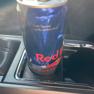 Op de afbeelding: Een blauwe en zilveren blikje Red Bull Energy Drink in een bekerhouder in de auto. Op het blikje staat "With Taurine. Lightly carbonated." en "Red Bull Energy Drink".