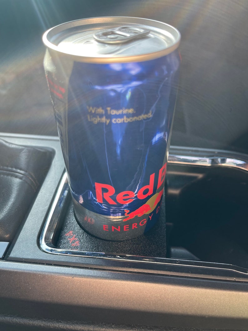 Op de afbeelding: Een blauwe en zilveren blikje Red Bull Energy Drink in een bekerhouder in de auto. Op het blikje staat "With Taurine. Lightly carbonated." en "Red Bull Energy Drink".