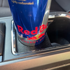 Op de afbeelding: Een blauwe en zilveren blikje Red Bull Energy Drink in een bekerhouder in de auto. Het blikje heeft de tekst "Red Bull Energy Drink" en "With Taurine, lightly carbonated" erop.