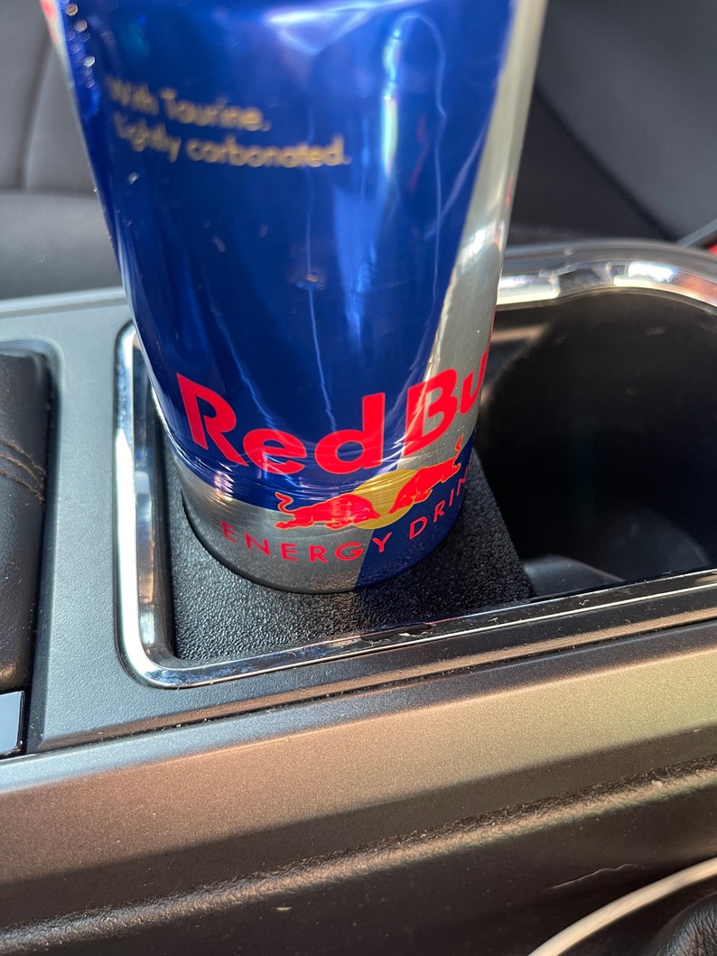 Op de afbeelding: Een blauwe en zilveren blikje Red Bull Energy Drink in een bekerhouder in de auto. Het blikje heeft de tekst "Red Bull Energy Drink" en "With Taurine, lightly carbonated" erop.