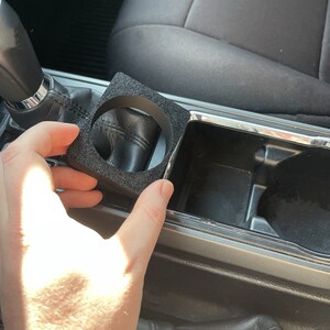 Op de afbeelding: Een zwarte schuimrubberen bekerhouderinzet voor een auto. De inzet is ontworpen om in een standaard bekerhouder te passen en helpt te voorkomen dat bekers rammelen of morsen.