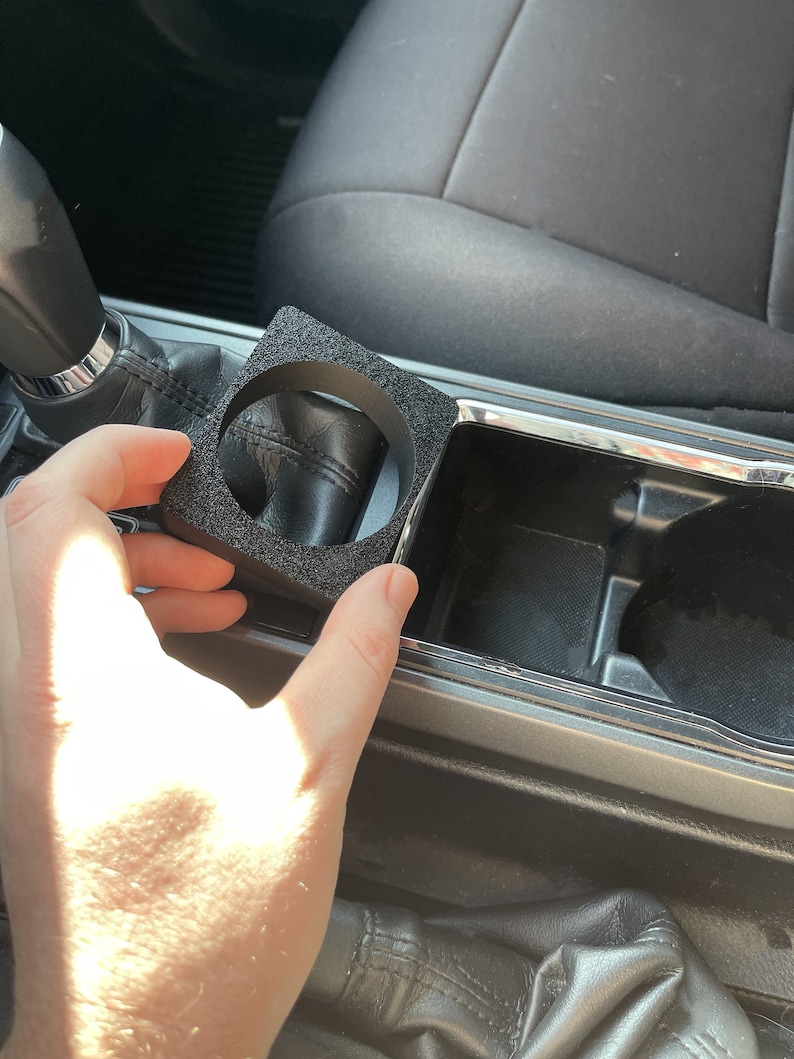 Op de afbeelding: Een zwarte schuimrubberen bekerhouderinzet voor een auto. De inzet is ontworpen om in een standaard bekerhouder te passen en helpt te voorkomen dat bekers rammelen of morsen.