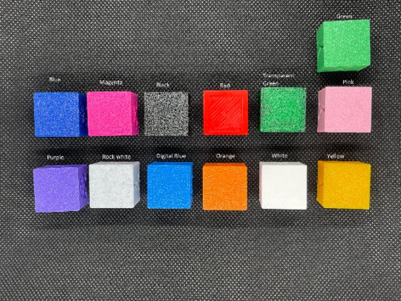 Op de afbeelding: Een verzameling van 12 kleine, vierkante, getextureerde blokken in verschillende kleuren, waaronder blauw, groen, rood, oranje, geel, roze, paars, wit, zwart en grijs. De blokken zijn gerangschikt in twee rijen van zes, waarbij elk blok is gelabeld met de bijbehorende kleur.