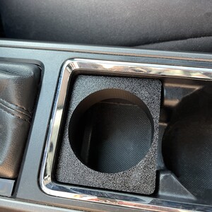 Op de afbeelding: Een zwarte bekerhouderinzet voor een auto. De inzet is gemaakt van een getextureerd materiaal en heeft een ronde opening voor een beker.