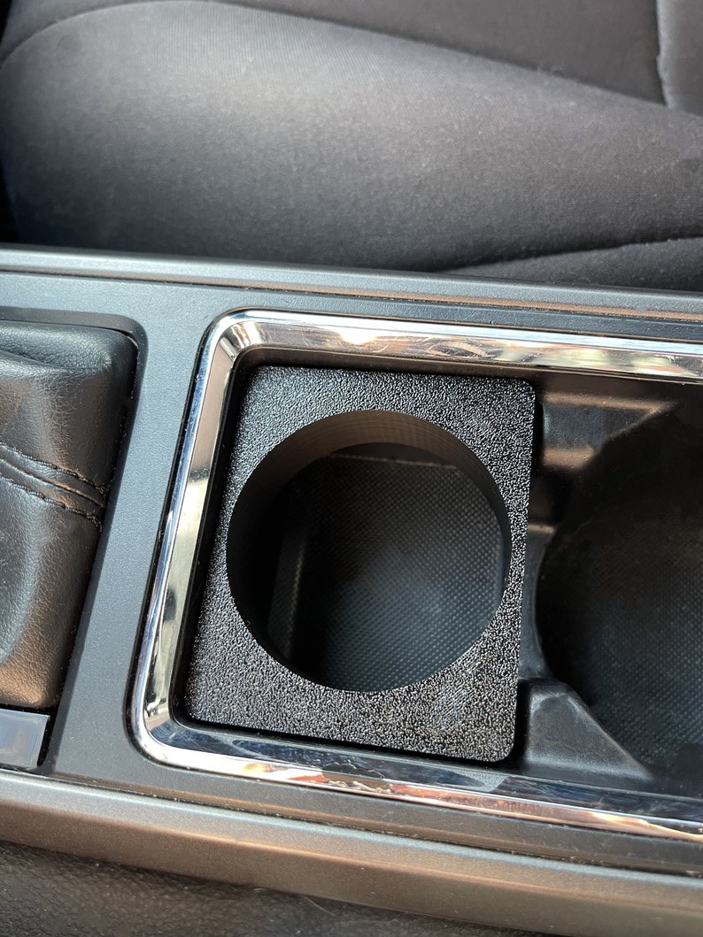 Op de afbeelding: Een zwarte bekerhouderinzet voor een auto. De inzet is gemaakt van een getextureerd materiaal en heeft een ronde opening voor een beker.
