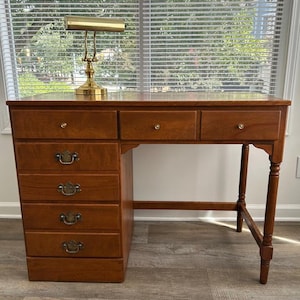 May include: A wooden desk with a medium brown finish featuring a left-side stack of four drawers with dark metal pulls and three top drawers with small round knobs. A brass-colored banker lamp with a rectangular shade sits on the desk surface.