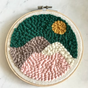 Può includere: Un cerchio da ricamo circolare con un design astratto colorato. Il design presenta un paesaggio verde, marrone, bianco e rosa con un sole giallo.