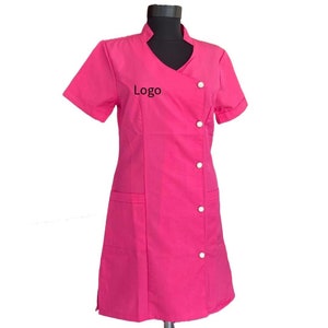 May include: A pink uniform dress with short sleeves and a V-neck collar. The dress has a button closure on the left side and a pocket on each side. The text "Logo" is embroidered on the front of the dress.