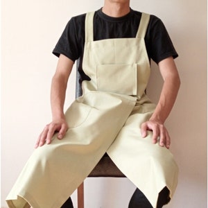 Może przedstawiać: Jasnozielony fartuch z dwoma szelkami i dużą kieszenią. Fartuch jest noszony przez osobę siedzącą na krześle.