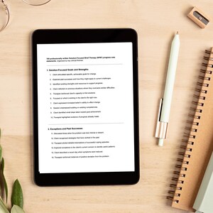 K&ouml;nnte beinhalten: Ein schwarzes Tablet mit Text zu Fortschrittsnotizen der l&ouml;sungsorientierten Kurztherapie (SFBT), zusammen mit einem wei&szlig;en Stift, einem Spiralnotizbuch und einem Anspitzer auf einer Holzoberfl&auml;che. Der Text enth&auml;lt nummerierte Listen.