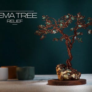Op de afbeelding: Een koperdraad boomsculptuur met groene edelsteen bladeren, gemonteerd op een rotsbasis en een houten standaard. De tekst "NEMA TREE RELIEF" wordt in het wit weergegeven. De achtergrond is donkerblauwgroen.