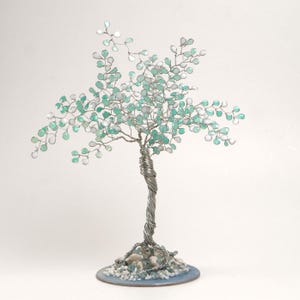Può includere: Una scultura artigianale di un albero in filo di ferro con un tronco argentato attorcigliato e rami adornati con perline di vetro verde acqua e trasparenti. L'albero poggia su una base circolare blu con piccole pietre.