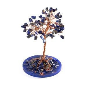 Op de afbeelding: Een handgemaakte boomsculptuur met een koperdraadstam en takken, versierd met blauwe en grijze edelsteenbladeren. De boom staat op een ronde, blauwe basis met bijpassende edelstenen.