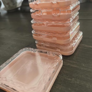 Könnte beinhalten: Ein Stapel aus sechs quadratischen, durchsichtigen Plastikboxen mit einem pfirsichfarbenen Innenraum. Die Boxen haben ein strukturiertes Design und sind übereinander gestapelt. Eine Box liegt offen auf einer Holzoberfläche.