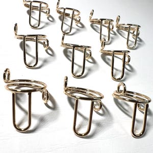 Puede incluir: Una colección de soportes para anillos de metal dorado. Cada soporte presenta una parte superior circular, un soporte en forma de U y un pequeño lazo en el lateral. Los soportes están dispuestos sobre una superficie blanca.