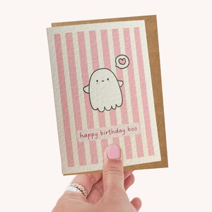 May include: A greeting card with a pink and white striped background. The card features a cute ghost illustration with a heart and the text "happy birthday boo". The card is held by a person.