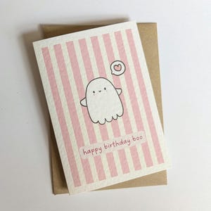 May include: A greeting card with a pink and white striped background, featuring a cartoon ghost with a heart above it. The text "happy birthday boo" is printed at the bottom. The card is on a brown envelope.