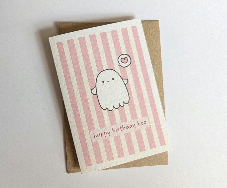 May include: A greeting card with a pink and white striped background, featuring a cartoon ghost with a heart above it. The text "happy birthday boo" is printed at the bottom. The card is on a brown envelope.