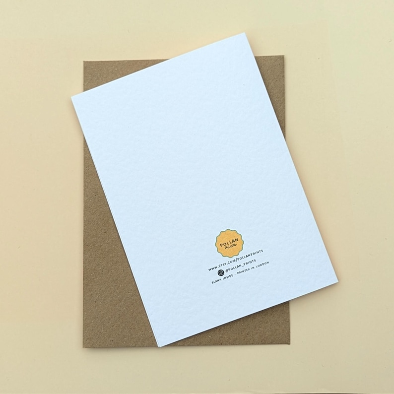 May include: A white greeting card with a textured surface, resting on a brown envelope. The card features a small logo and text at the bottom. The background is a light beige color.