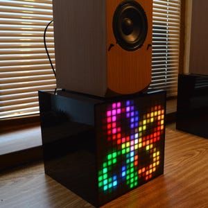 May include: Two wooden speakers sit atop black, cube-shaped stands with colorful LED displays. The speakers have a light wood finish and dark speaker cones. The LED displays show abstract, geometric patterns in various colors.