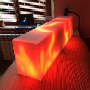 May include: A long, rectangular, white light box with a red and orange glow emanating from inside. The light box is sitting on a red surface.