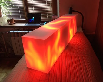 May include: A long, rectangular, white light box with a red and orange glow emanating from inside. The light box is sitting on a red surface.