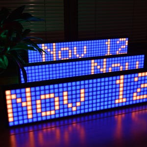 Może przedstawiać: Trzy niebieskie wyświetlacze LED ułożone jeden na drugim. Wyświetlacze pokazują tekst "Now 12" w pomarańczowych i białych światełkach.