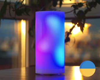 May include: A white cylindrical lamp with a colorful, abstract design. The lamp is lit and emits a blue, purple, and pink glow. The lamp is sitting on a wooden table.