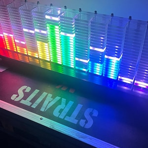 May include: A colorful LED sound visualizer with clear acrylic columns. The display cycles through a spectrum of colors, including red, orange, yellow, green, and blue. The word "STRAITS" is stenciled on the base.