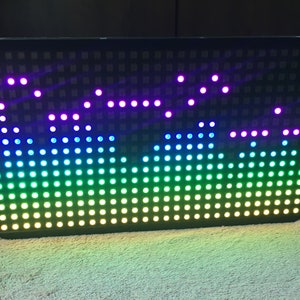 Può includere: Un pannello LED rettangolare nero con una griglia di luci che mostra uno spettacolo di luci colorate. Le luci sono disposte a forma di onda e cambiano colore dal viola al blu, dal verde al giallo.