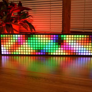 Peut inclure: Un écran LED rectangulaire avec un cadre noir, affichant un ensemble vibrant de carrés colorés. L'écran est éclairé d'un spectre de couleurs, dont le vert, le rose, le bleu et le jaune. L'écran repose sur une surface en bois, reflétant la lumière colorée.
