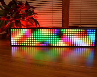 Puede incluir: Una pantalla LED rectangular con un marco negro, que muestra una vibrante matriz de cuadrados coloridos. La pantalla está iluminada con un espectro de colores, incluyendo verde, rosa, azul y amarillo. La pantalla se encuentra sobre una superficie de madera, reflejando la luz colorida.