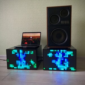 May include: Two black cube-shaped speakers with a blue and green pixelated design on the front. A laptop computer is sitting on top of one of the speakers. A large black speaker with a wooden frame is sitting on top of the other speaker.